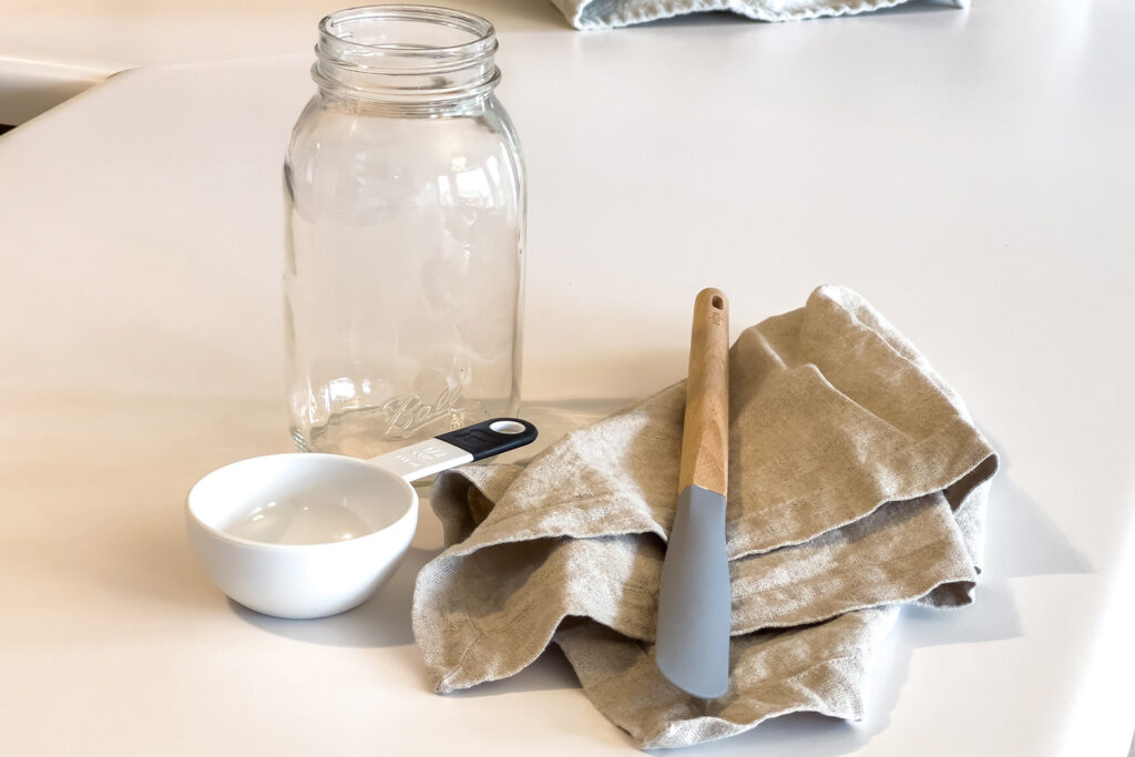 a jar, a measuring cup, a spatula, and a linen cloth sit on the counter, for why sourdough starter
