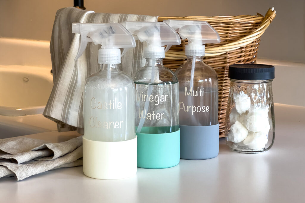 a jar of diy garbage disposal cleaning pods, a bottle of Castile Cleaner Spray, a bottle of vinegar cleaning spray, and a bottle of multi purpose cleaning spray sit next to each other