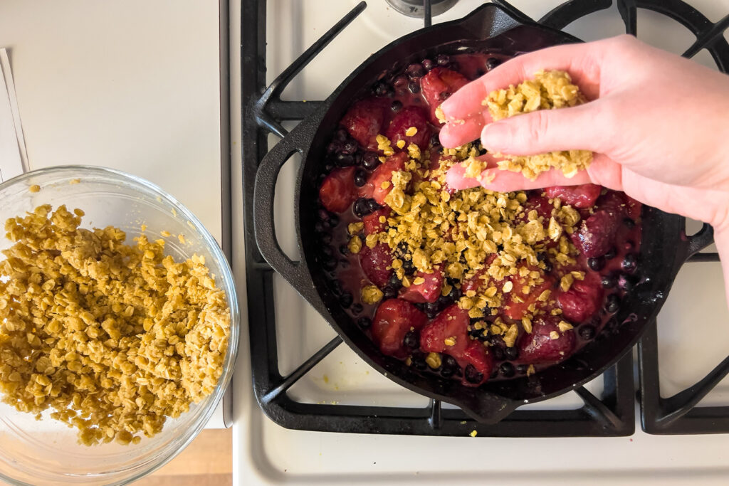 a woman is sprinkling crumble topping on top of the berry filling in a cast iron skillet