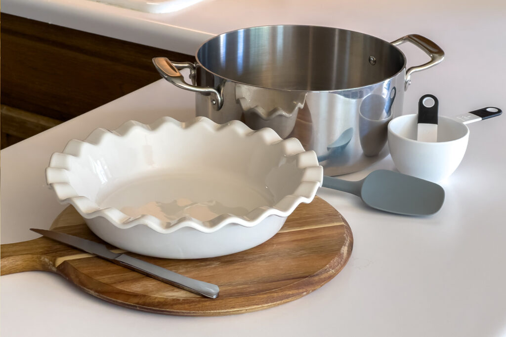 a pie plate, a pot, a measuring cup and spoon, a spatula, and a cutting board and knife sit on the kitchen counter