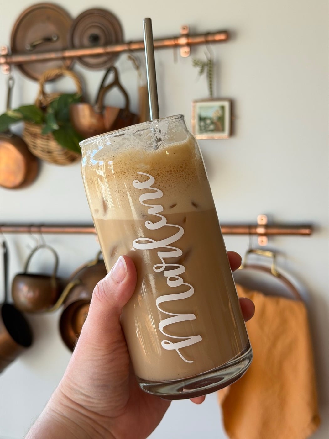 Marlene holding her iced salted maple latte in a glass with her name on it, copper pots in the background