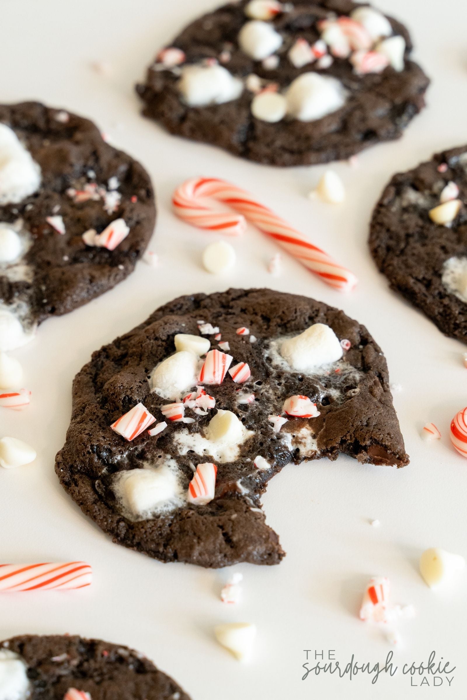 Peppermint hot cocoa cookies with white chocolate chips, marshmallows, and candy canes, one with a bite taken out.