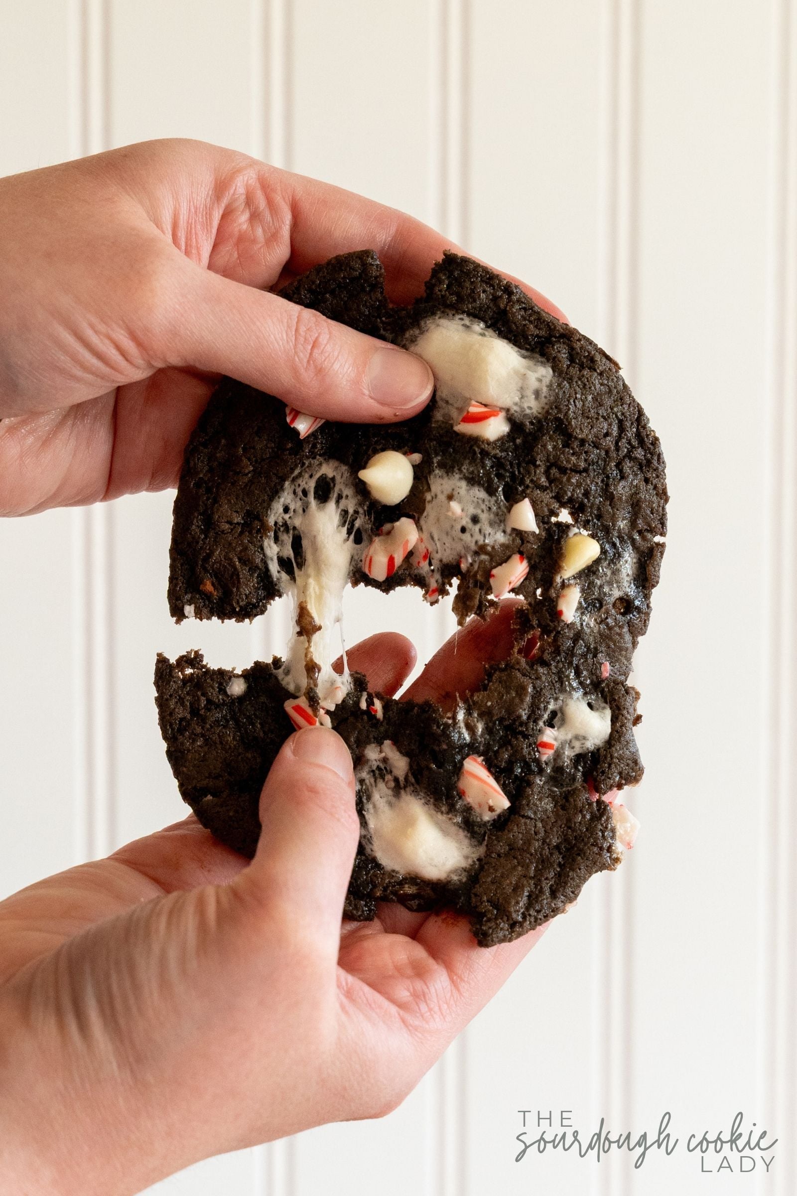 Hands pulling apart a sourdough peppermint hot cocoa cookie to show its gooey marshmallow center.
