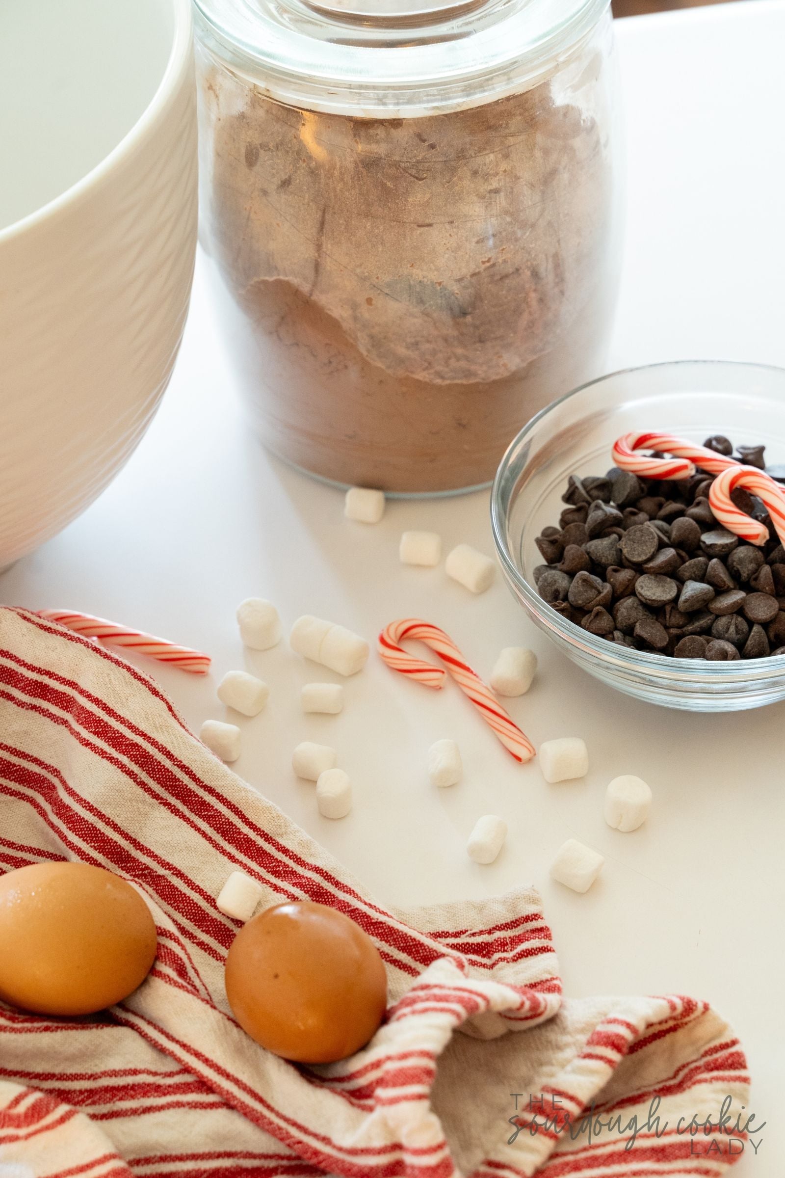 Ingredients for sourdough peppermint hot cocoa cookies, including cocoa powder, chocolate chips, mini marshmallows, candy canes, and eggs.